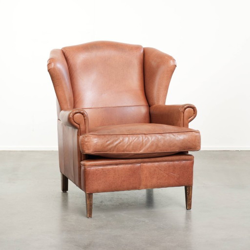 Fauteuil à oreilles en cuir de vachette couleur cognac clair, avec passepoil assorti et un style anglais plein de caractère.