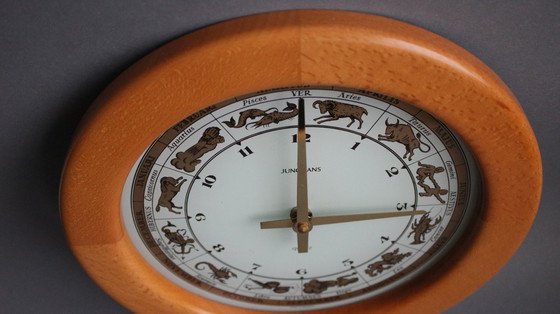 Image 1 of Horloge murale vintage Junghans avec boîtier en bois et signe du zodiaque sur un cadran en verre.
