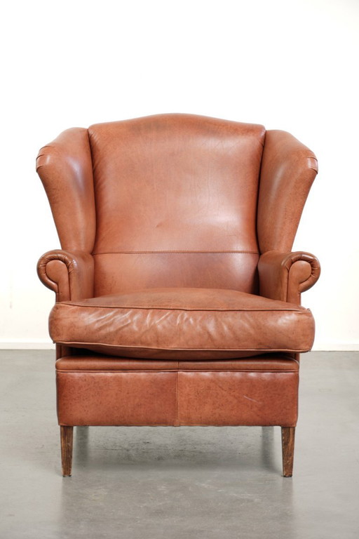 Fauteuil à oreilles en cuir de vachette couleur cognac clair, avec passepoil assorti et un style anglais plein de caractère.